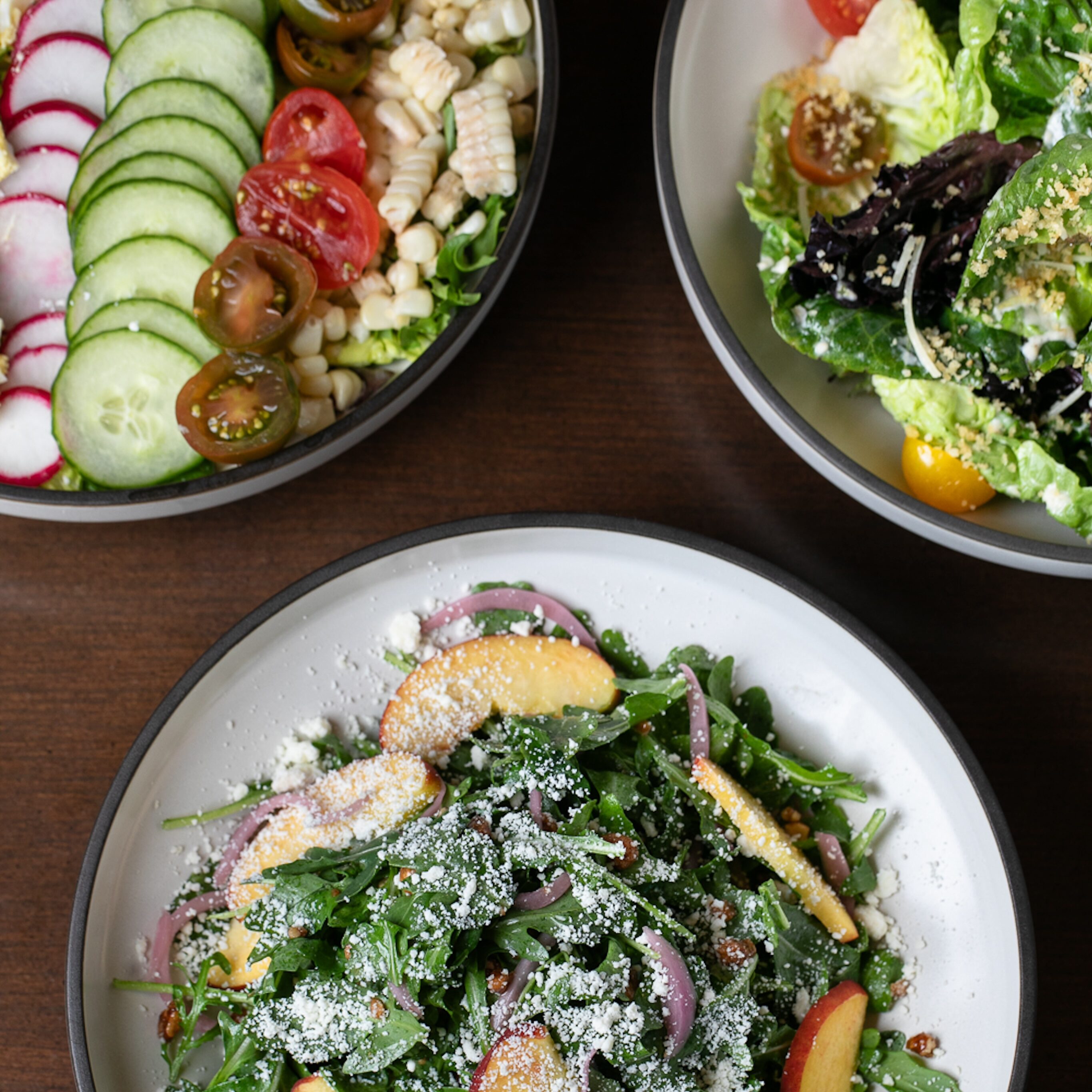 A gallery of flavor awaits with three bowls of salad adorning a wooden table: one featuring mixed greens and parmesan, another brimming with sliced cucumbers and corn, and a third offering assorted greens and tomatoes.