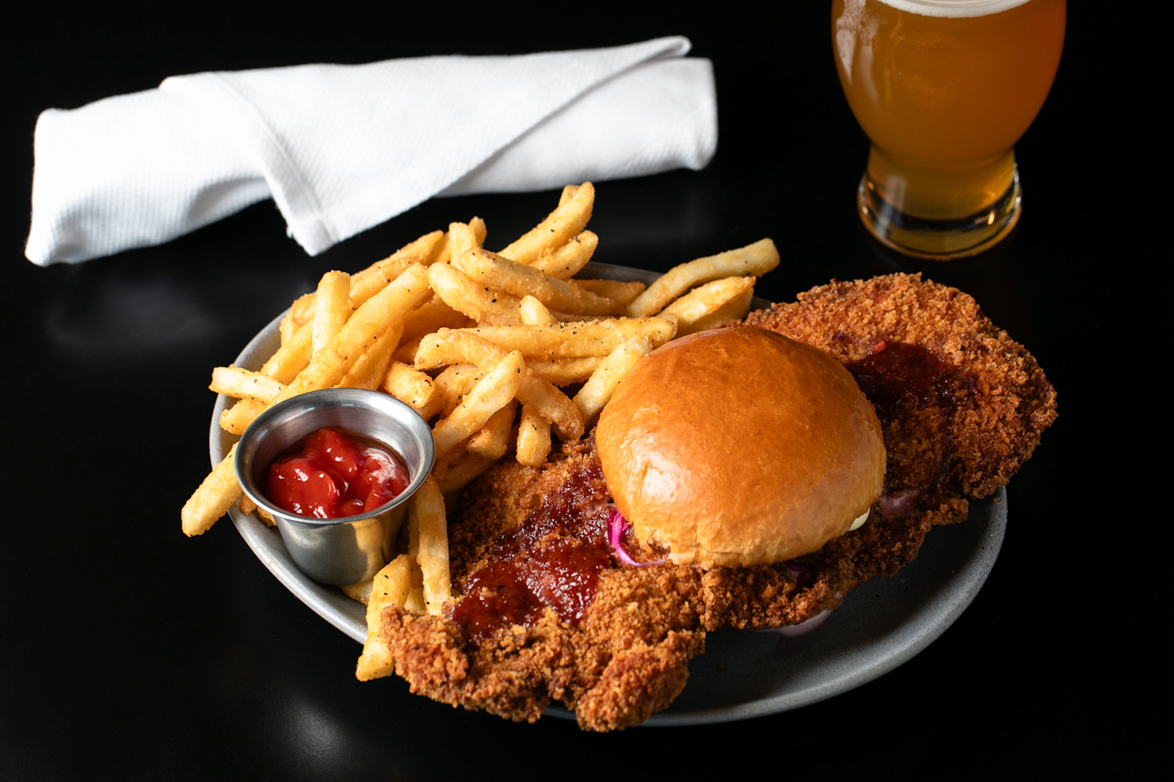 A gallery-worthy plate featuring a breaded chicken sandwich, crispy fries paired with ketchup, a neatly folded napkin, and a chilled glass of beer rests elegantly on the dark surface.