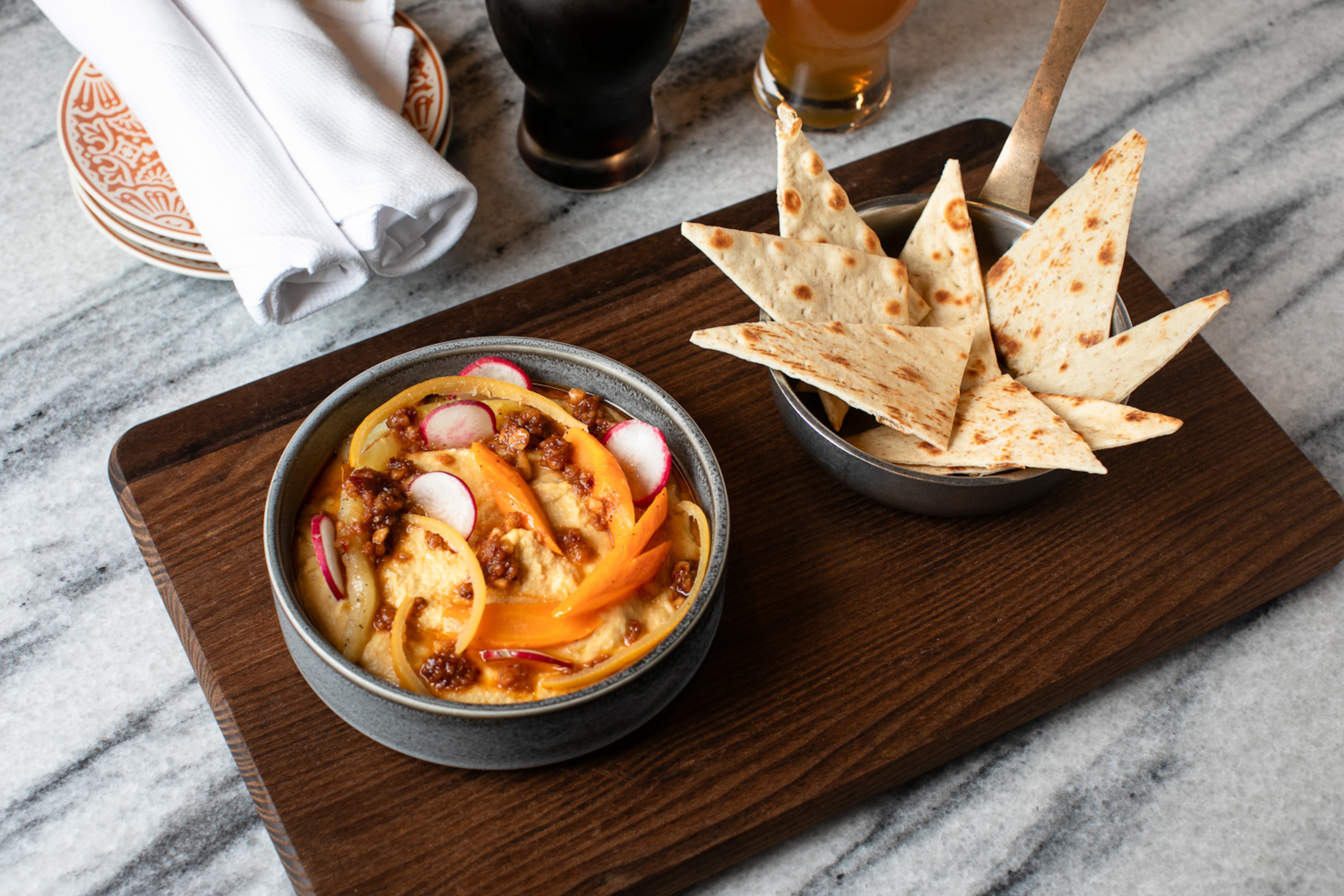 A bowl of dip garnished with sliced radishes and peppers, next to a bowl of triangular flatbreads, is displayed like an art piece in a culinary gallery on a wooden board.