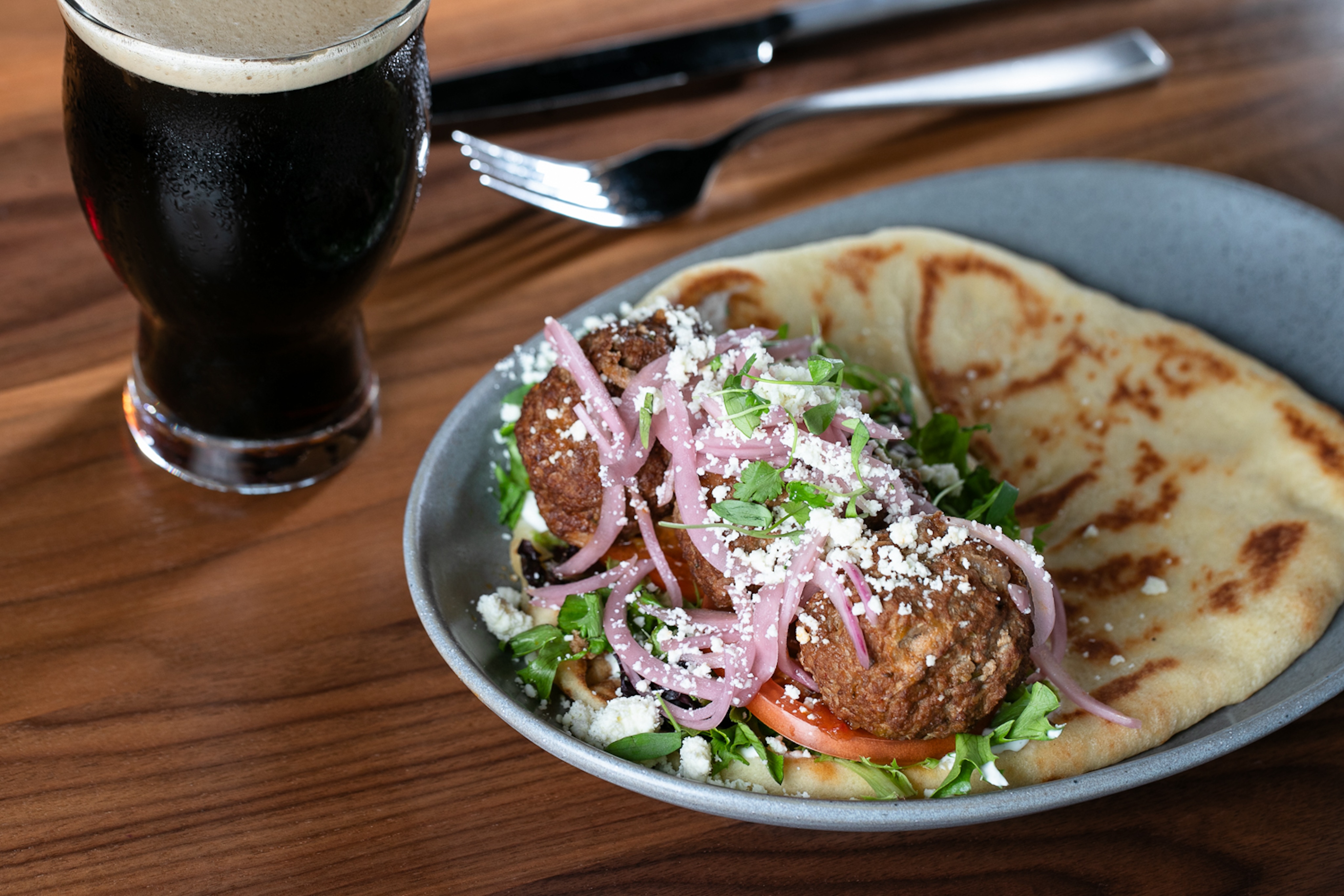 A plate with falafel, pickled onions, greens, and feta on flatbread sits like a masterpiece in a gallery next to a glass of dark beer on a wooden table.