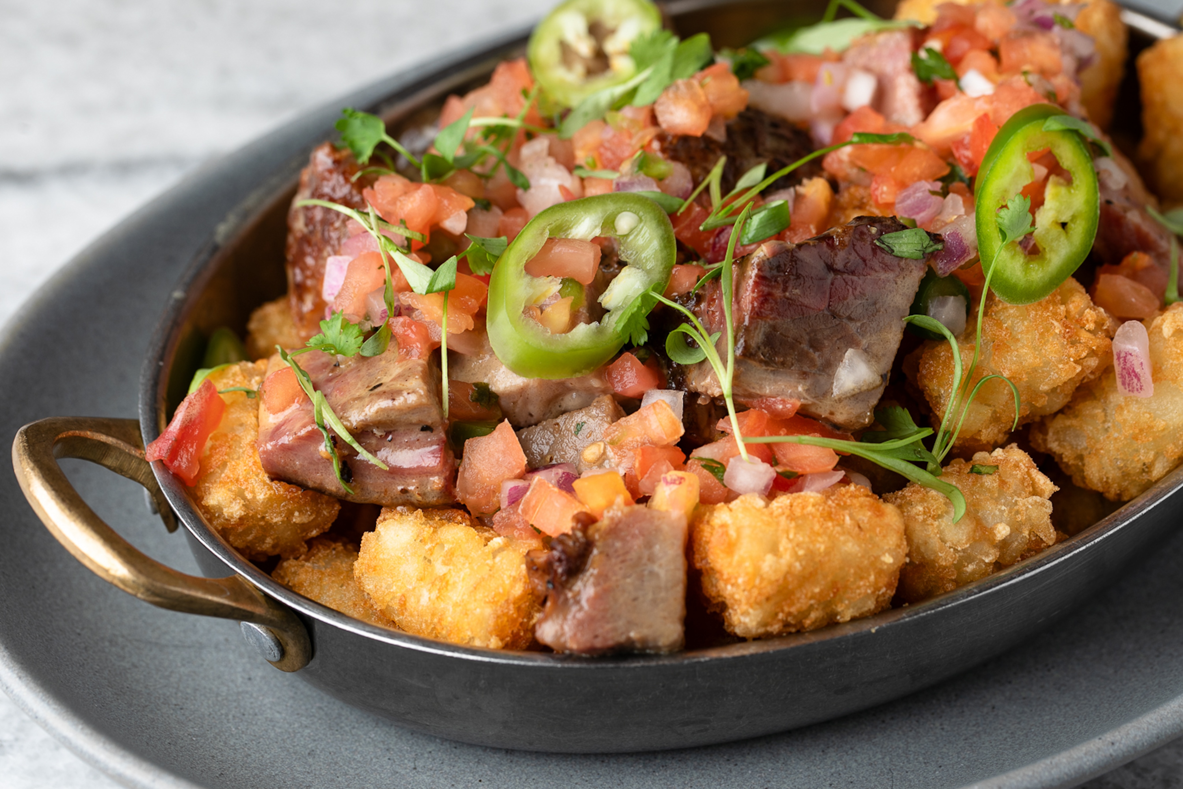 A dish of crispy tater tots topped with diced steak, chopped tomatoes, green jalapeño slices, red onions, and fresh herbs is a true culinary gallery presented in a gray metal bowl with handles.