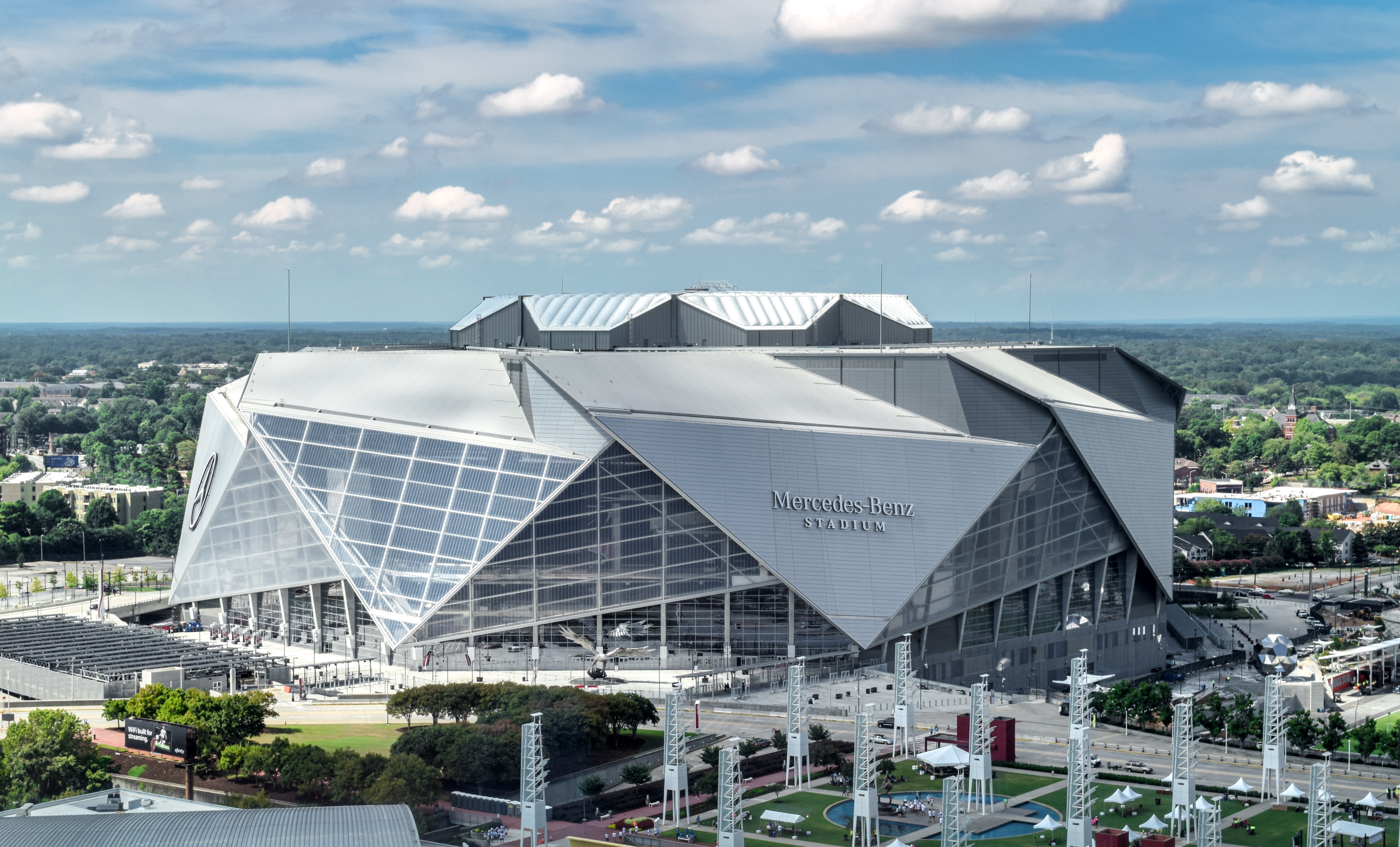 Aerial view of the large, geometrically-designed Mercedes-Benz Stadium with its iconic signage, set amidst a cityscape dotted with trees and nearby State Farm Arena, all under a partly cloudy sky.