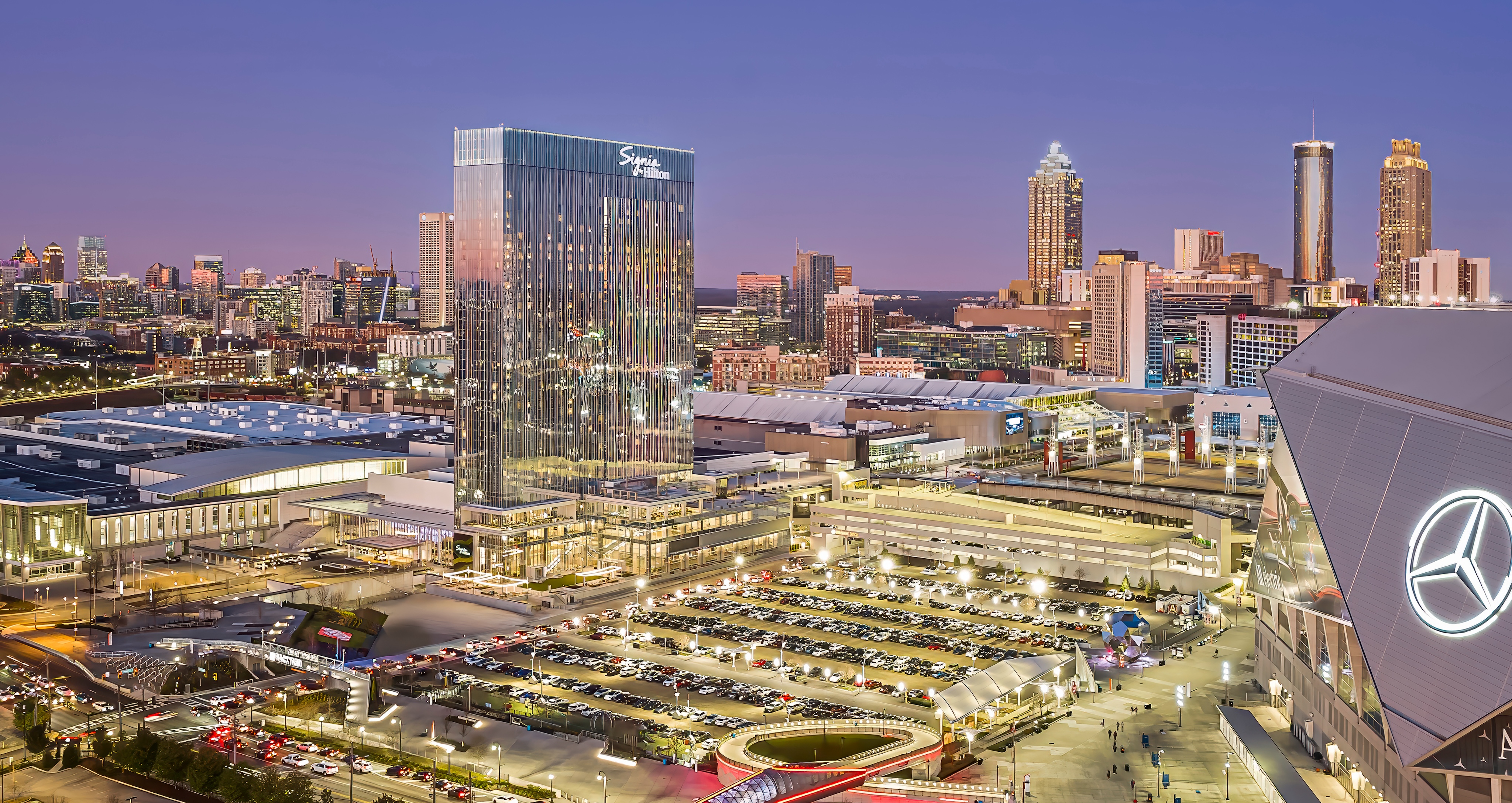 Aerial view of an illuminated urban skyline at dusk featuring tall buildings, a stadium with a Mercedes-Benz logo, and a bustling parking lot surrounded by popular attractions and restaurants.