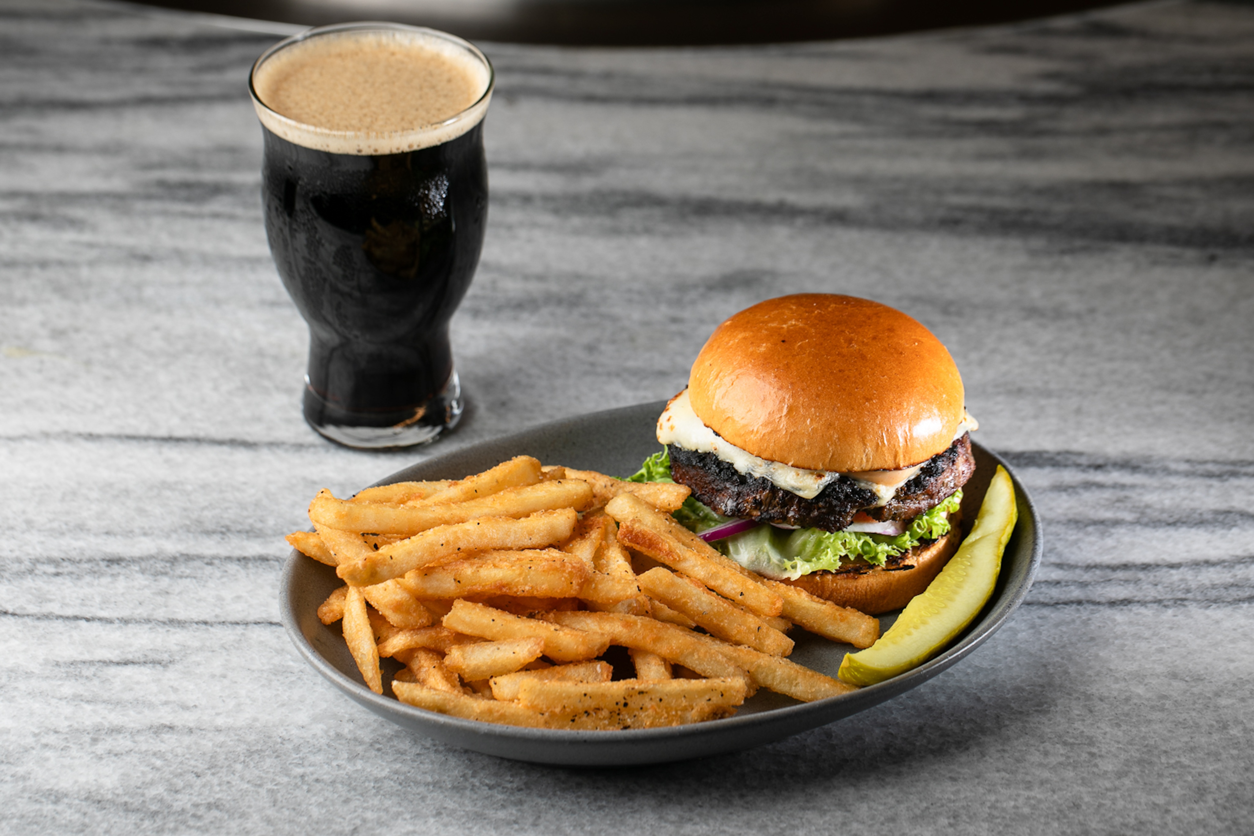 A glass of dark beer sits beside a burger, seasoned fries, and a pickle spear on a marble surface—an Instagram-worthy setup you'd find at one of Atlanta's best sports bars.