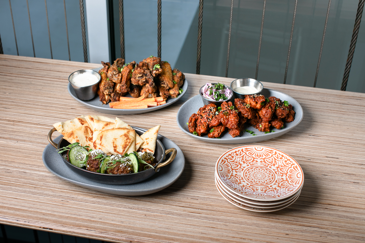 Three plates of food on a table: grilled skewers with pita, fried wings with fries and dip, and saucy cauliflower bites with dressing, alongside stacked patterned plates.