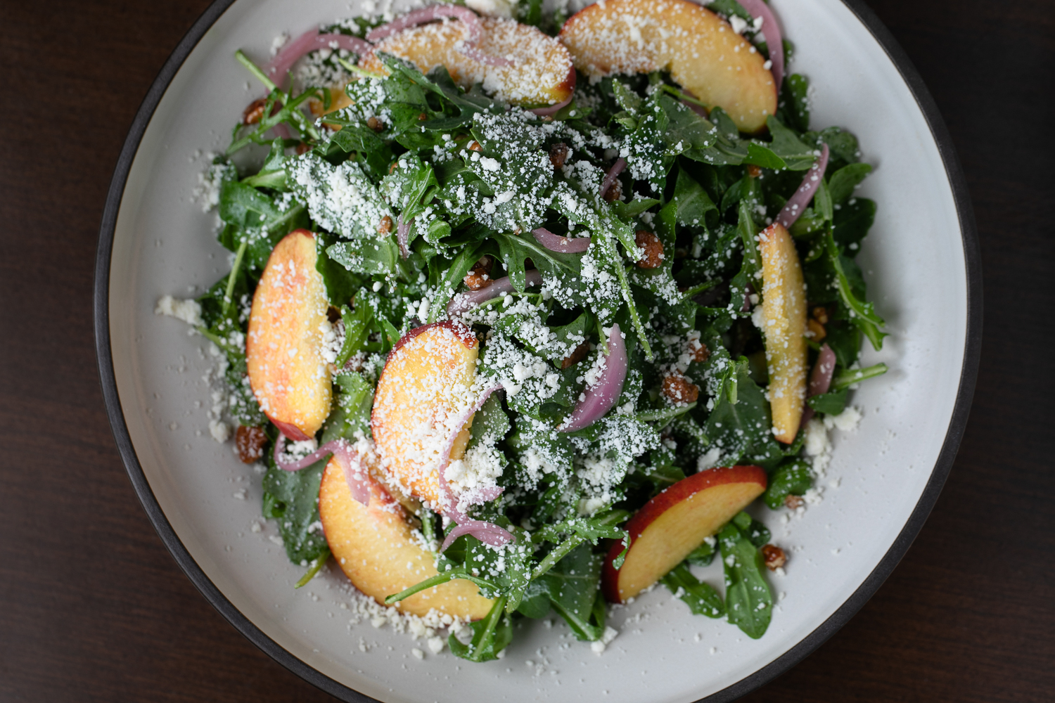 A salad artistically arranged like a gallery masterpiece features arugula, sliced peaches, red onions, raisins, and grated cheese on a white plate.