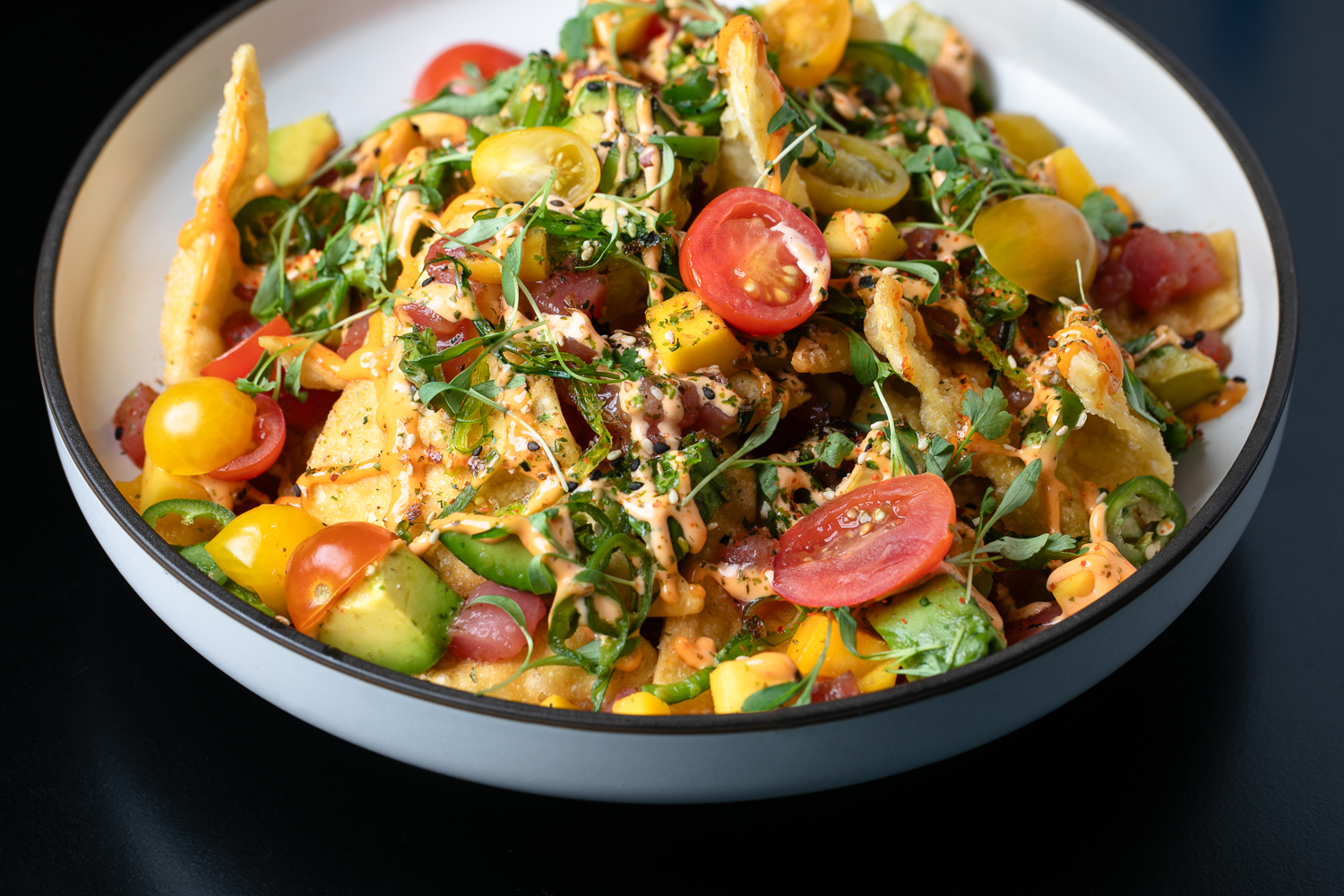 A vibrant gallery of flavors, this bowl of colorful nachos is artfully topped with cherry tomatoes, avocado, herbs, and a drizzle of sauce.