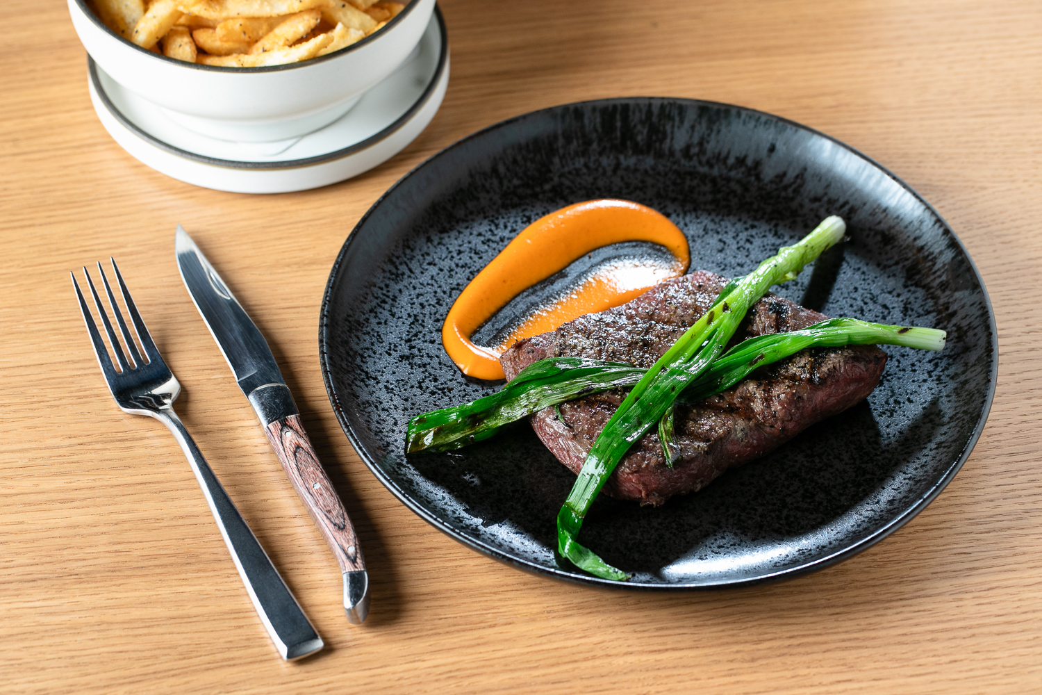 On a sleek black plate, a gallery-worthy presentation features steak garnished with green onions and orange sauce, complemented by a knife and fork. In the background, a bowl of fries subtly peeks into the scene.