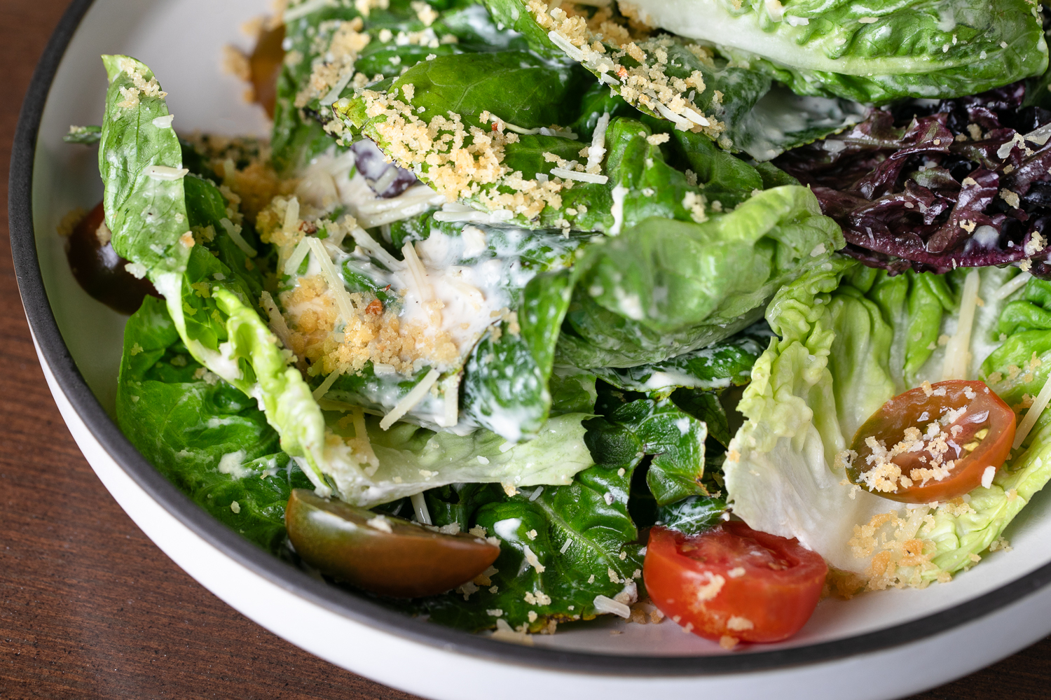 A bowl of Caesar salad artfully arranged like a gallery piece, featuring romaine lettuce, cherry tomatoes, grated cheese, and croutons.