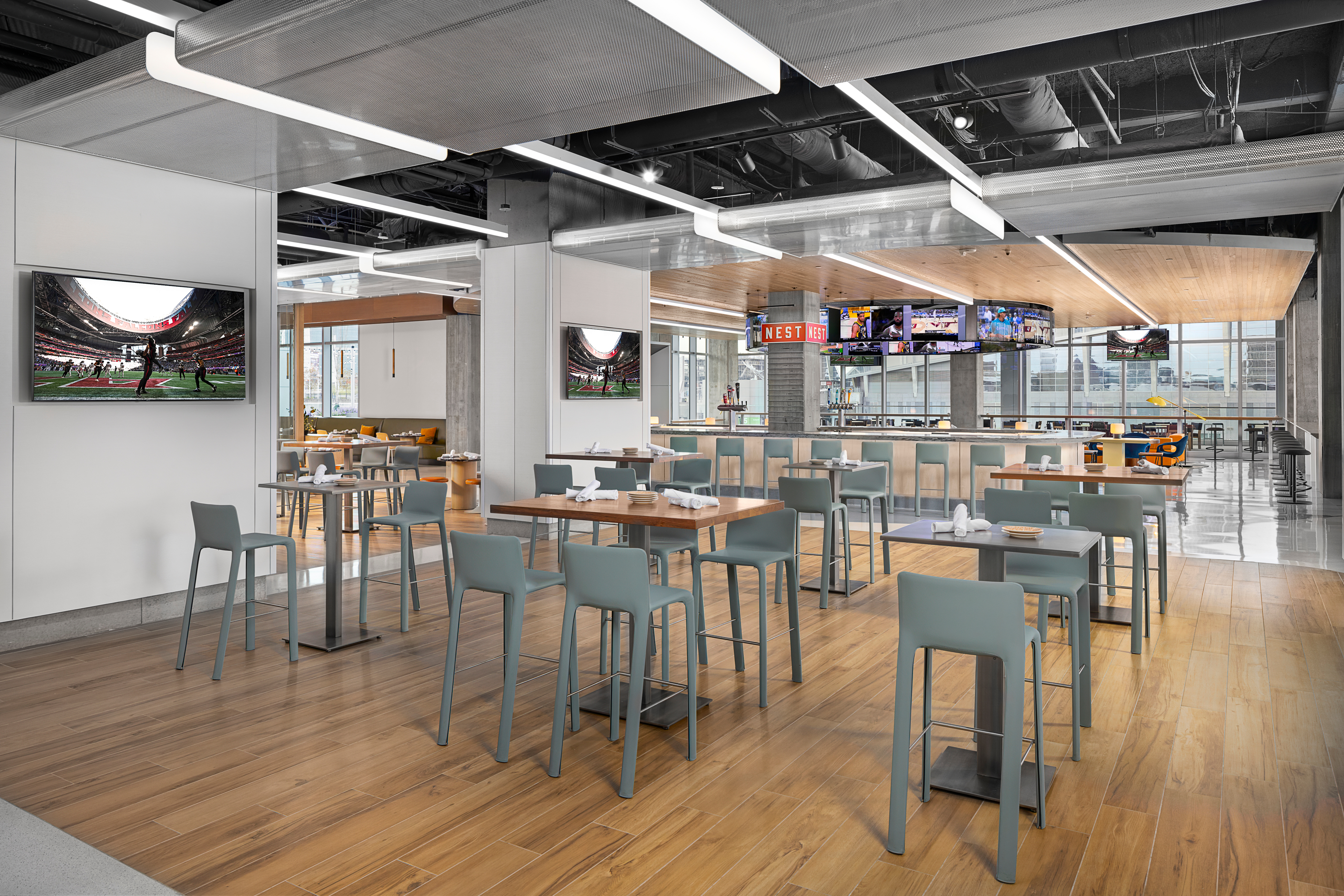 Modern dining area with wooden flooring, tables, chairs, and multiple TVs. Ceiling features exposed ducts, and large windows offer natural light.