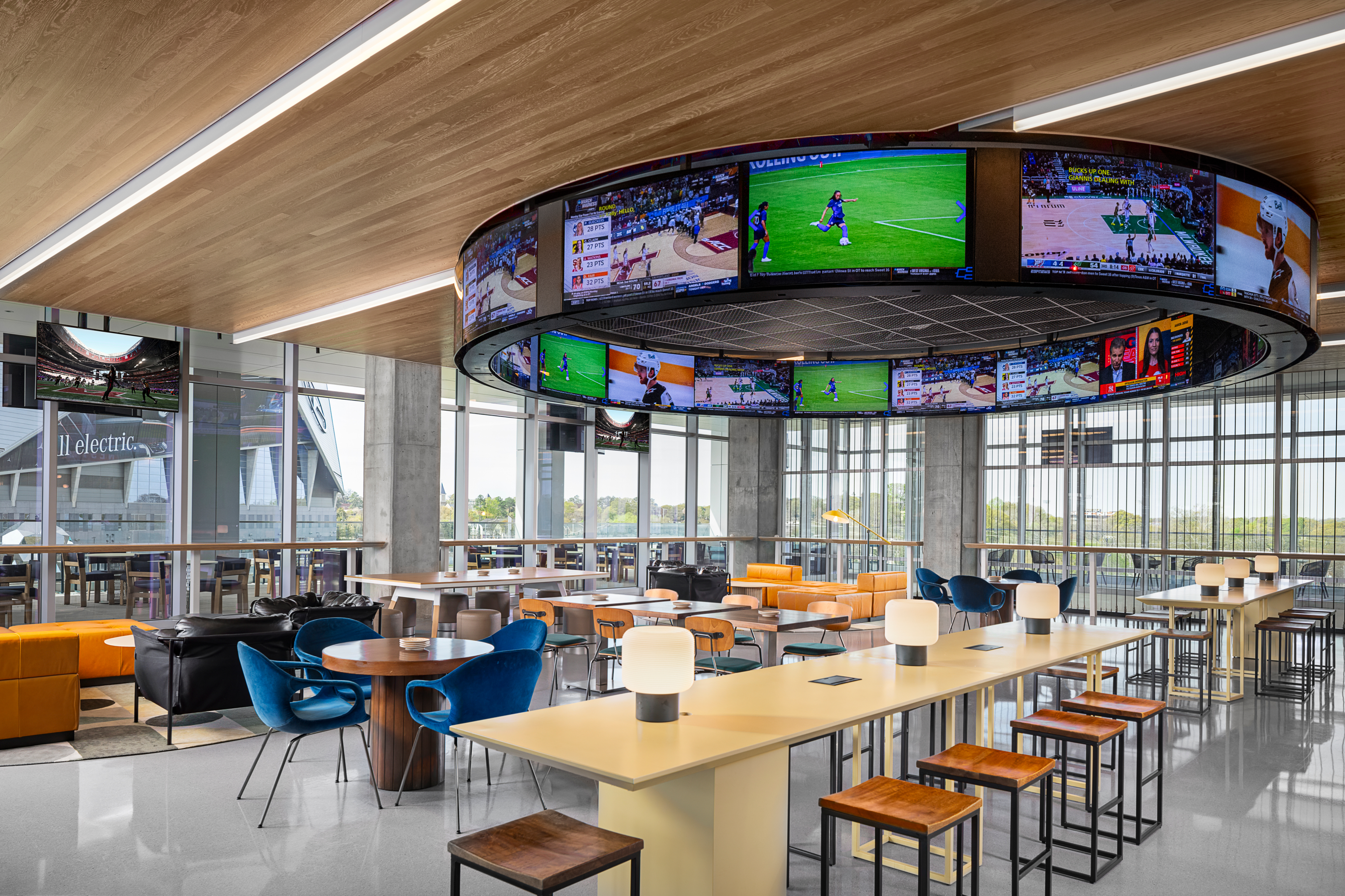 Modern sports bar with circular overhead screens, assorted seating, and wooden tables. Natural light streams through large windows.