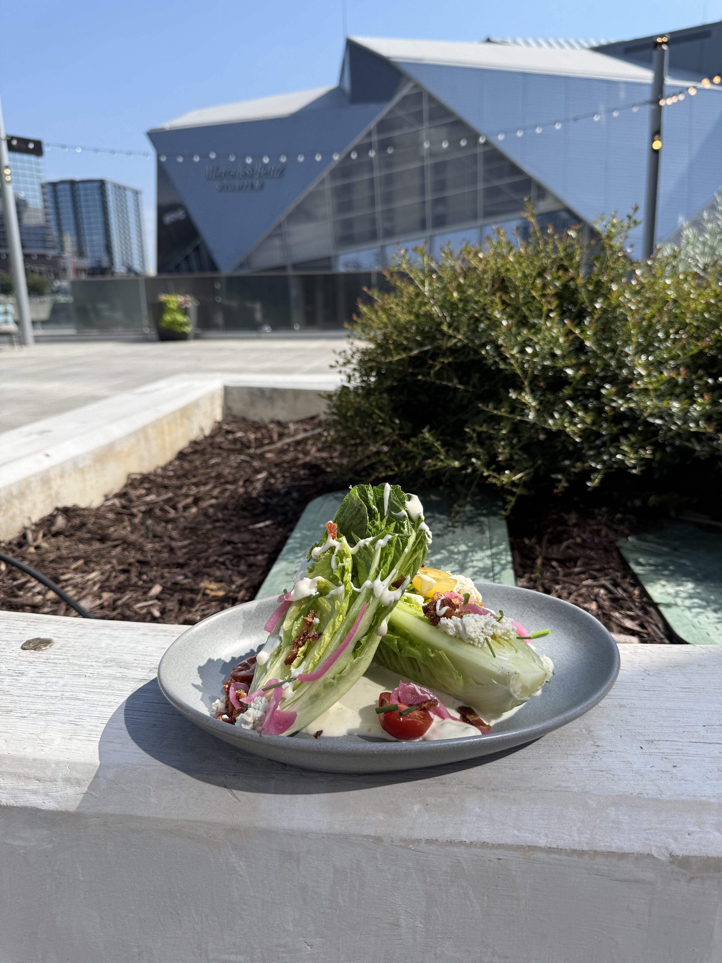 A wedge salad with tomatoes, onions, dressing, and greens on a gray plate, placed outdoors with a modern building in the background.