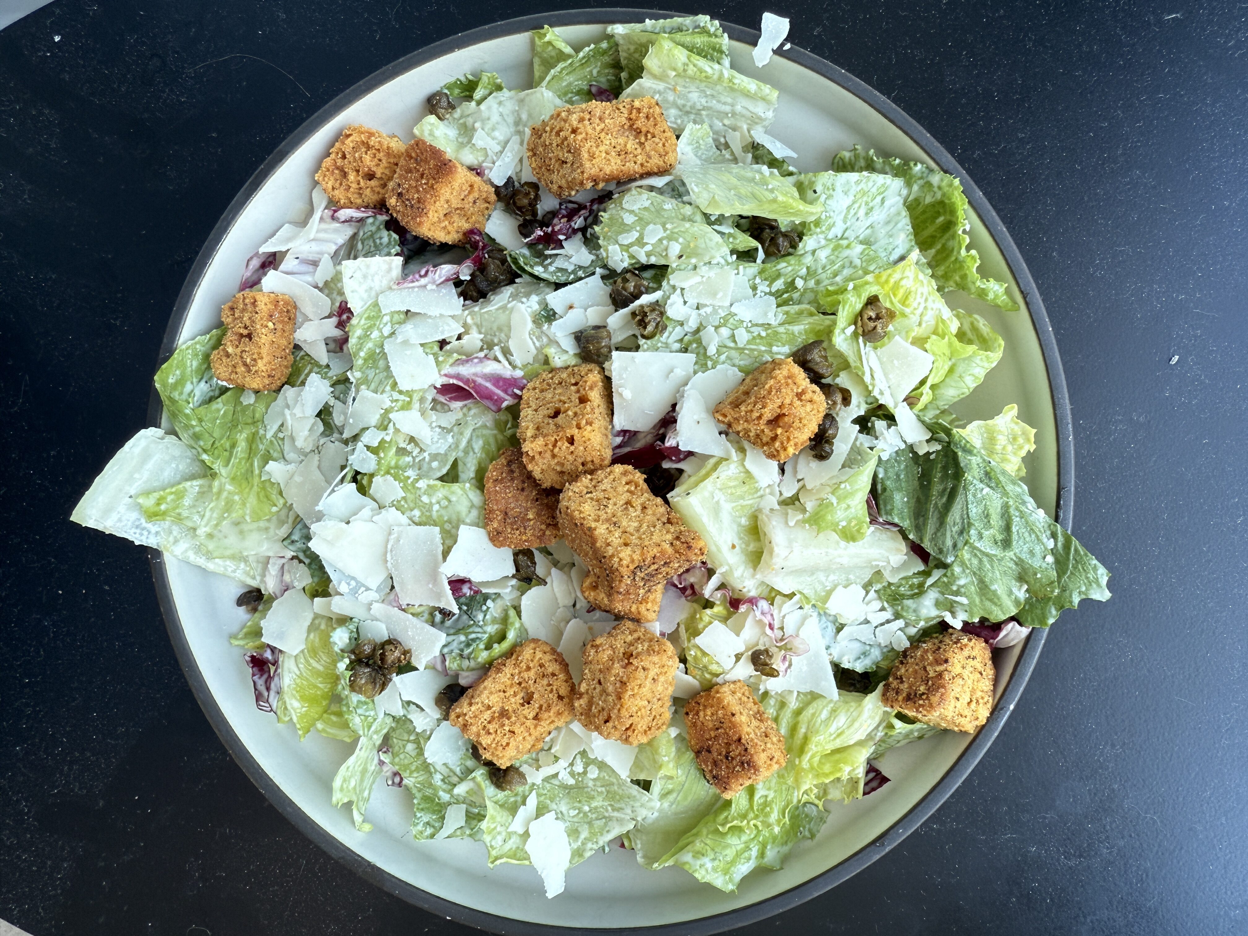 A plate of Caesar salad with romaine lettuce, shredded cheese, croutons, and capers on a dark surface.