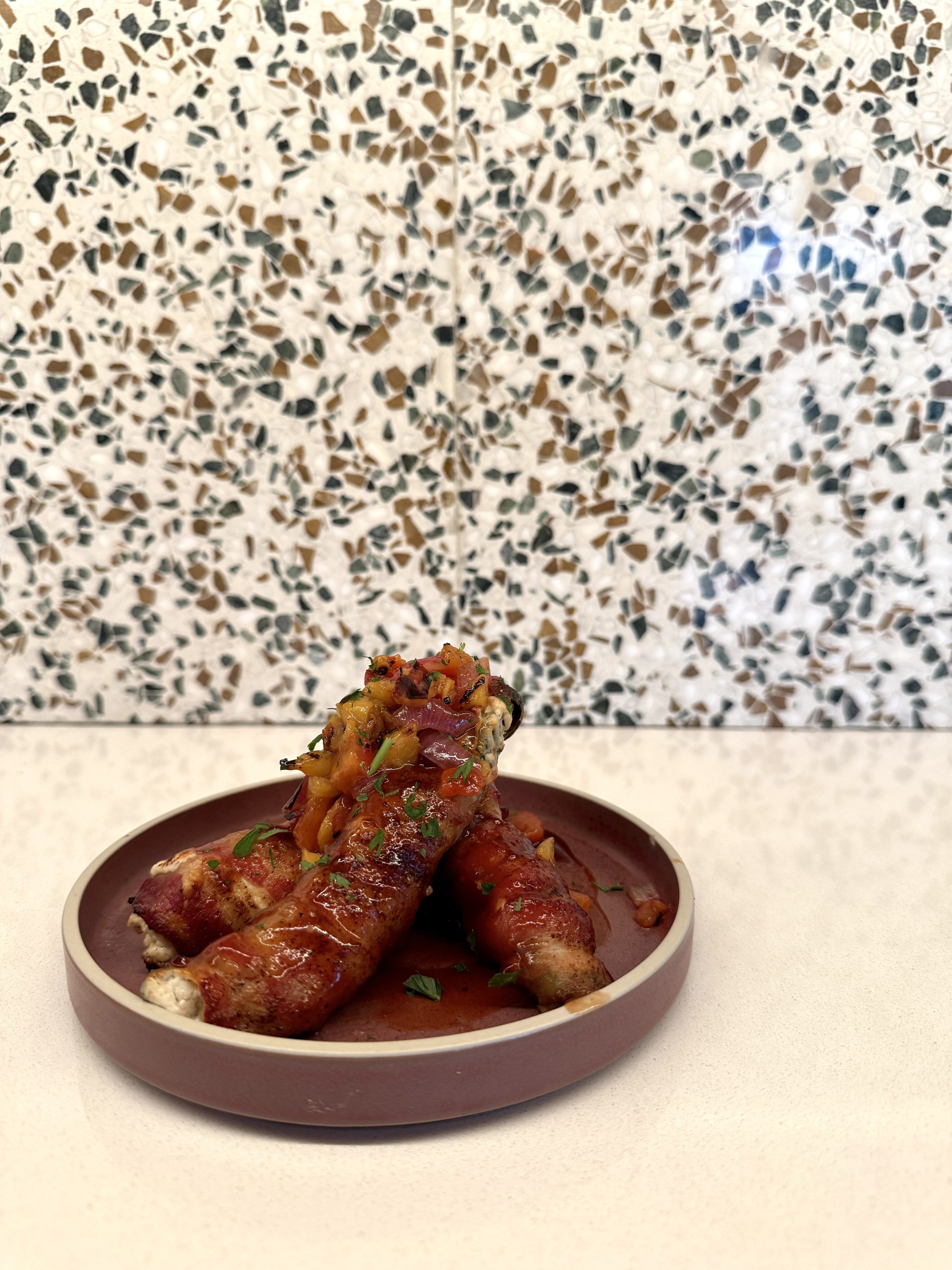 A round plate with three sausages topped with tomato-based sauce and vegetables, set on a white counter with a patterned tile background.