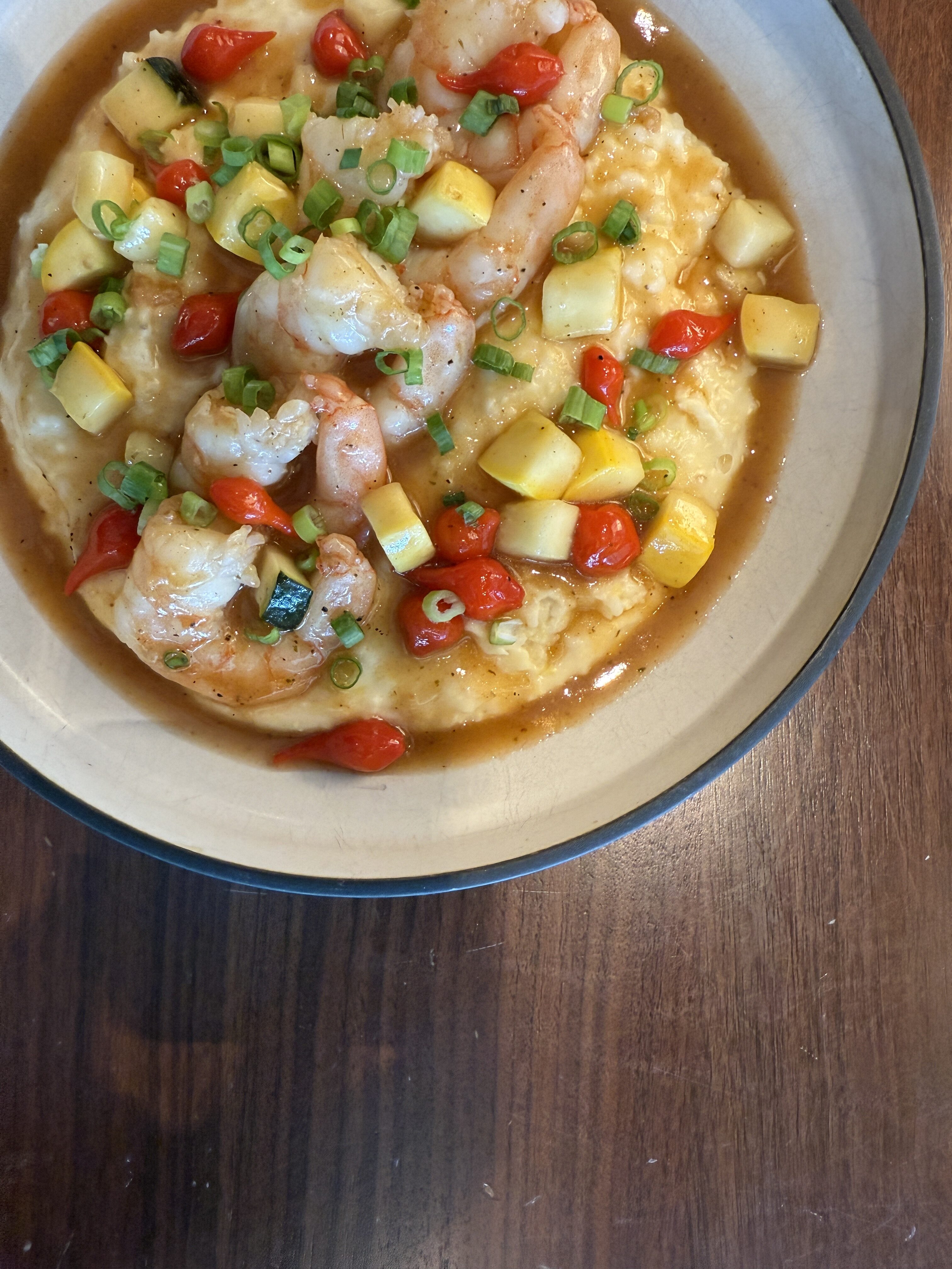 A bowl of shrimp and grits topped with diced yellow squash, cherry tomatoes, green onions, and a brown sauce, served on a wooden table.