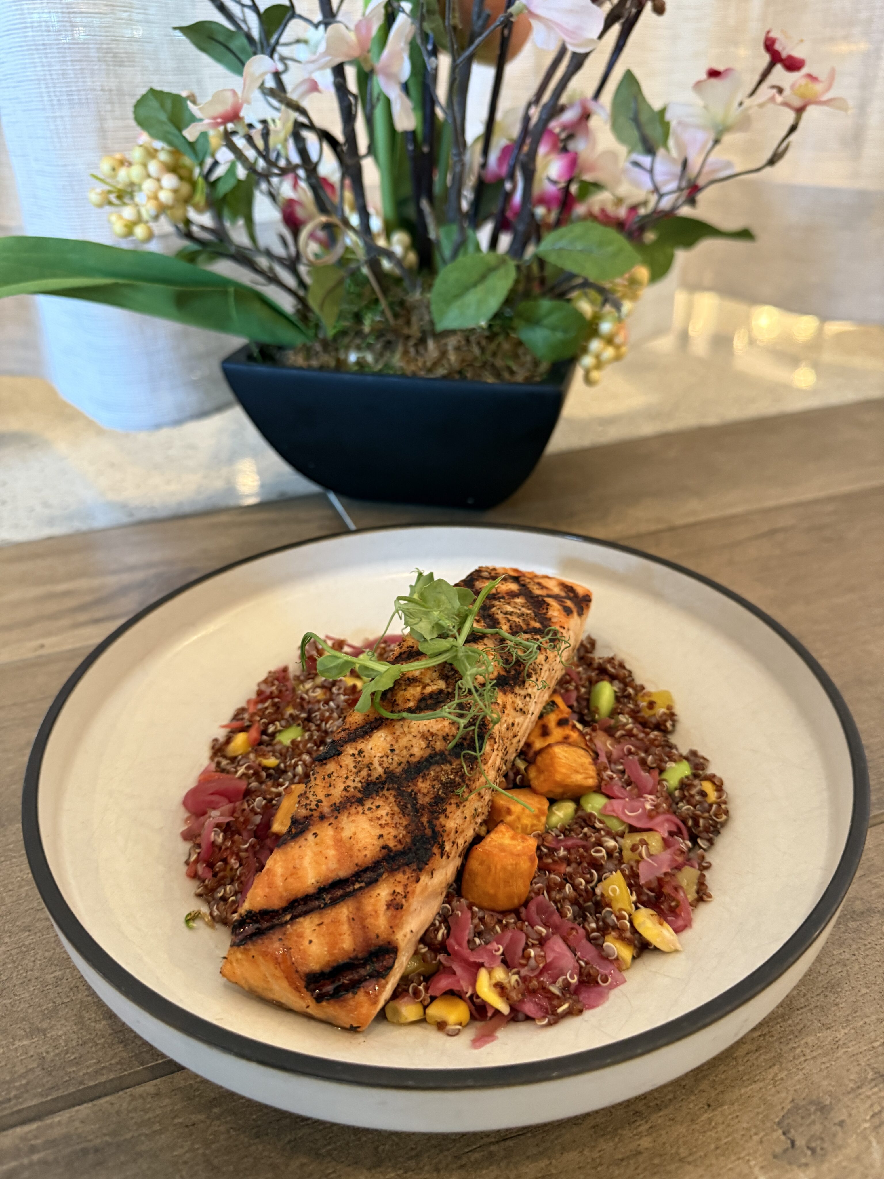 A grilled salmon fillet served on a bed of quinoa with vegetables, garnished with herbs, sits on a plate in front of a potted plant with flowers.
