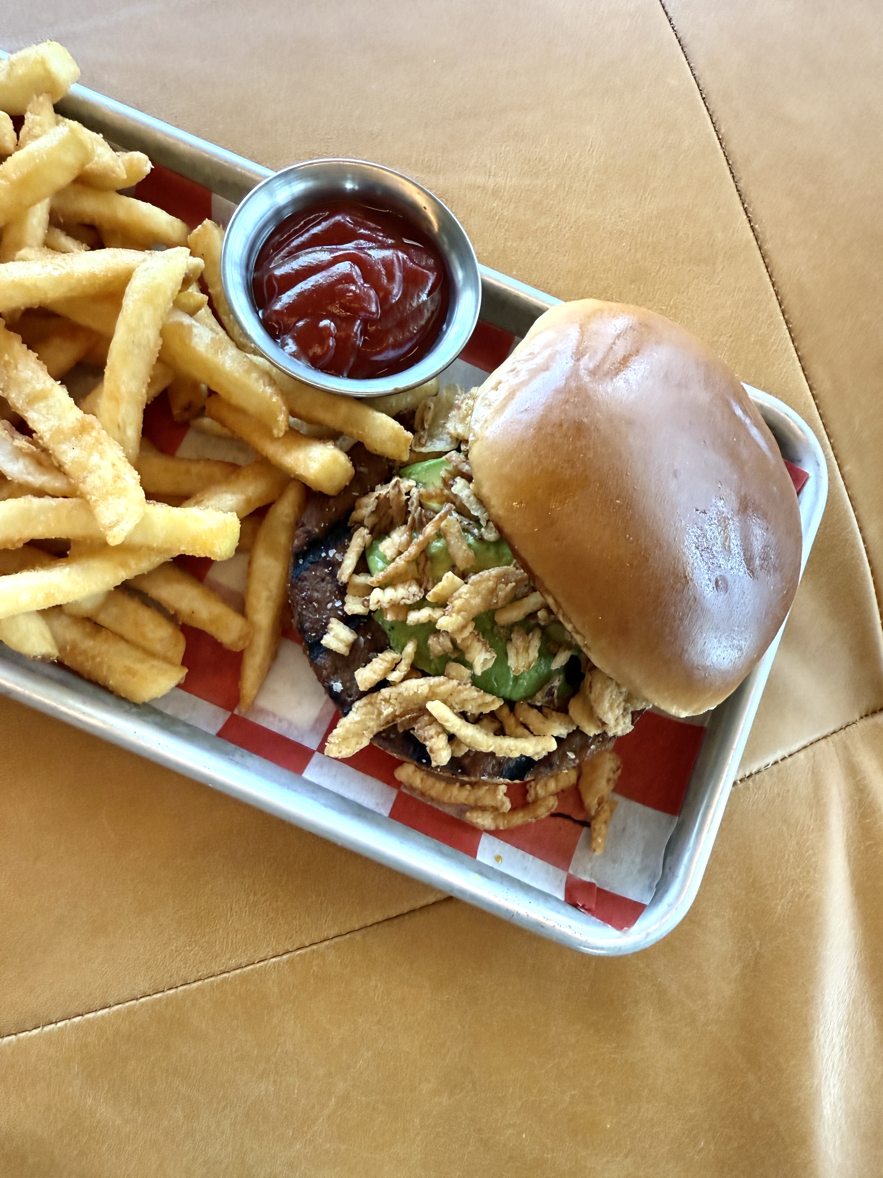 A tray with a burger topped with crispy onions and green garnish, a side of French fries, and a metal cup of ketchup, on a tan surface.