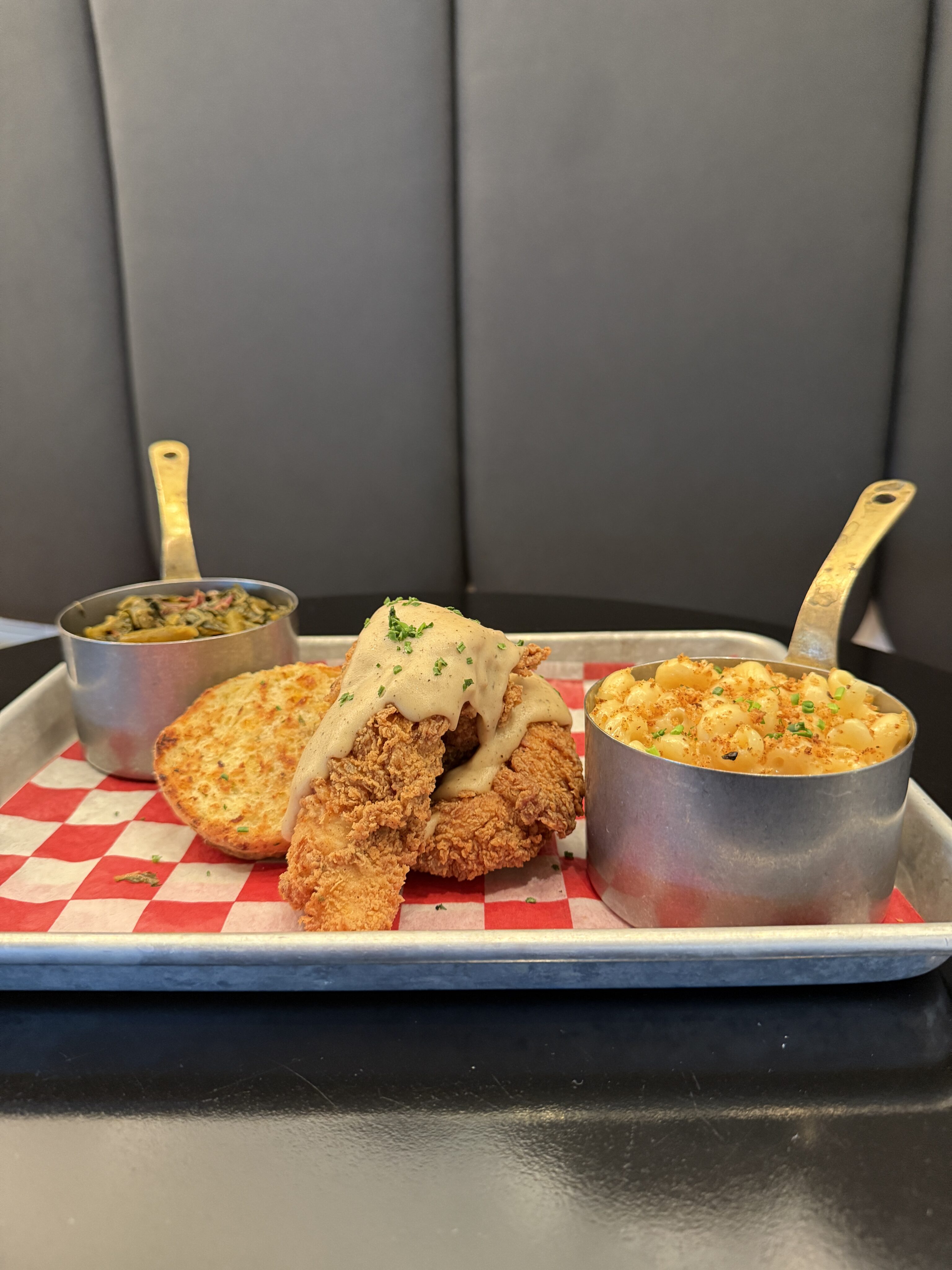 A metal tray with fried chicken topped with gravy, a biscuit, macaroni and cheese, and a cup of greens, served on a red and white checkered paper.