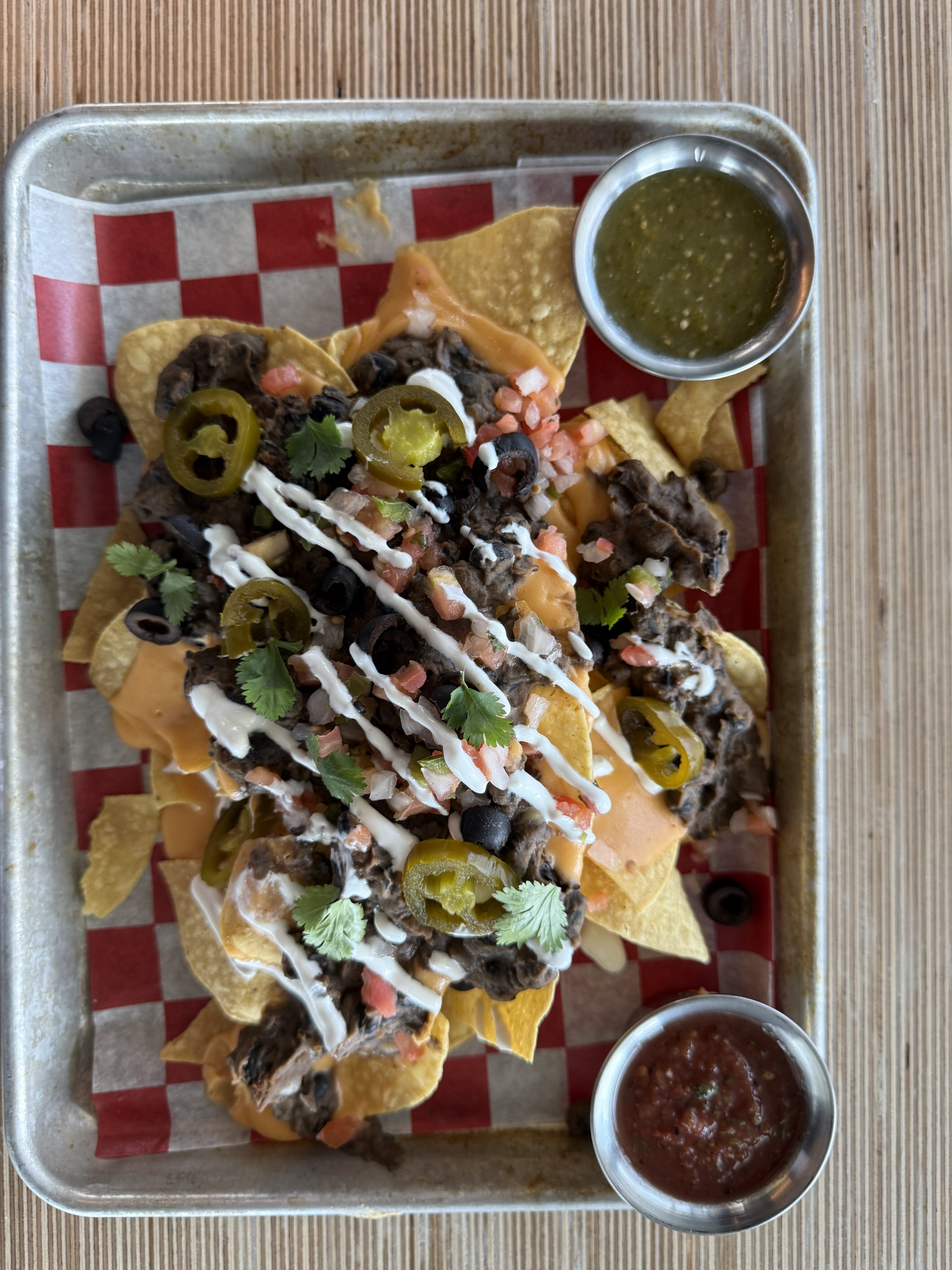 A tray of loaded nachos topped with beef, cheese, jalapeños, black olives, cilantro, and drizzled sauce, served with green and red salsa on the side.