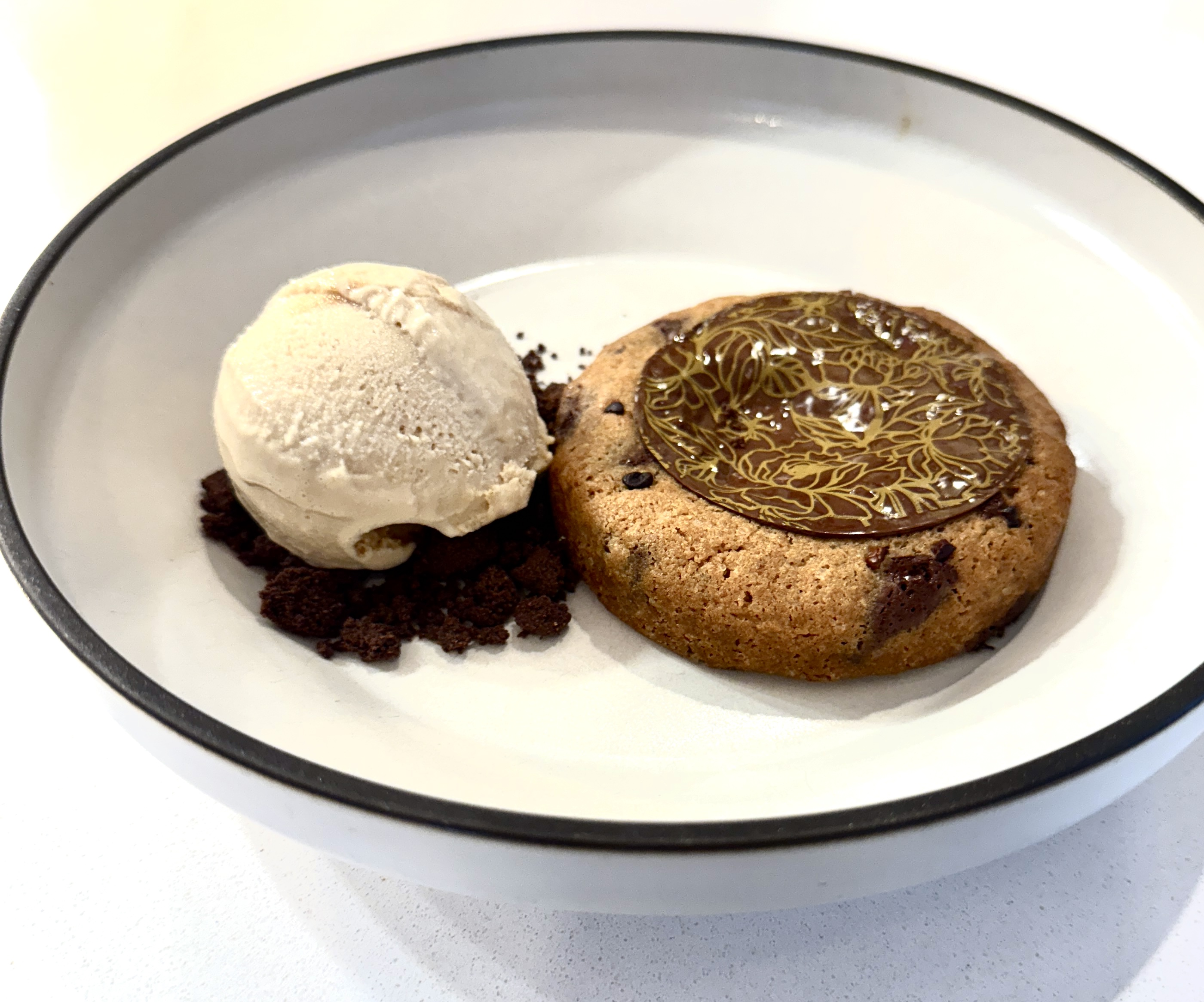 A scoop of ice cream on chocolate crumbs beside a chocolate chip cookie topped with glossy chocolate, served in a white bowl with a black rim.